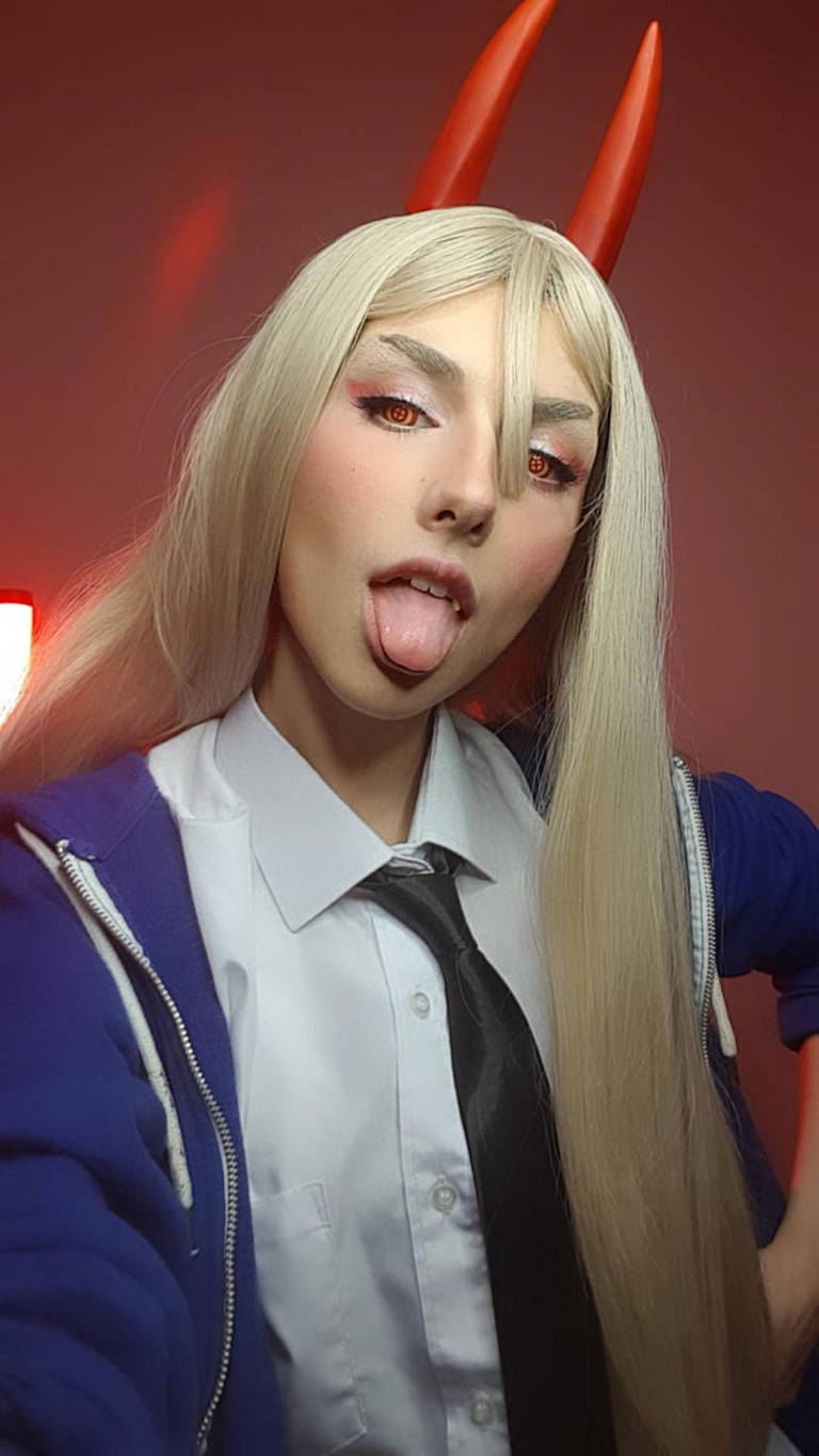 A person with long blonde hair and orange eyes sticking out their tongue, wearing a school uniform shirt, tie, and a blue hoodie with red horns on their head. The background is soft red lighting.