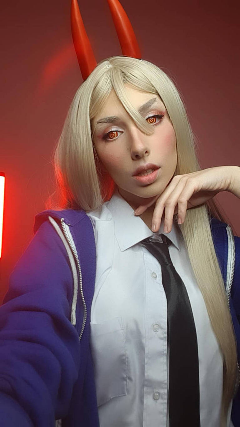 A person with long blonde hair and red horns, wearing a white shirt and black tie, poses with a hand on their chin against a dark background illuminated by red light.