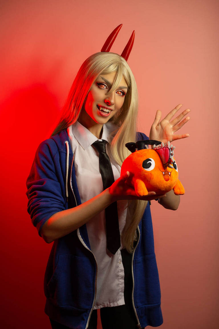 A person with long blonde hair and red horns is wearing a blue hoodie and a black tie, holding a plush toy. The background is pink with red lighting.