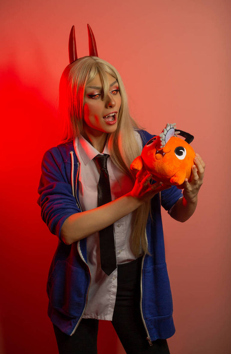 A person dressed in a blue hoodie and a white shirt with a black tie, holding an orange plush toy while expressing surprise or excitement, with red lighting in the background.