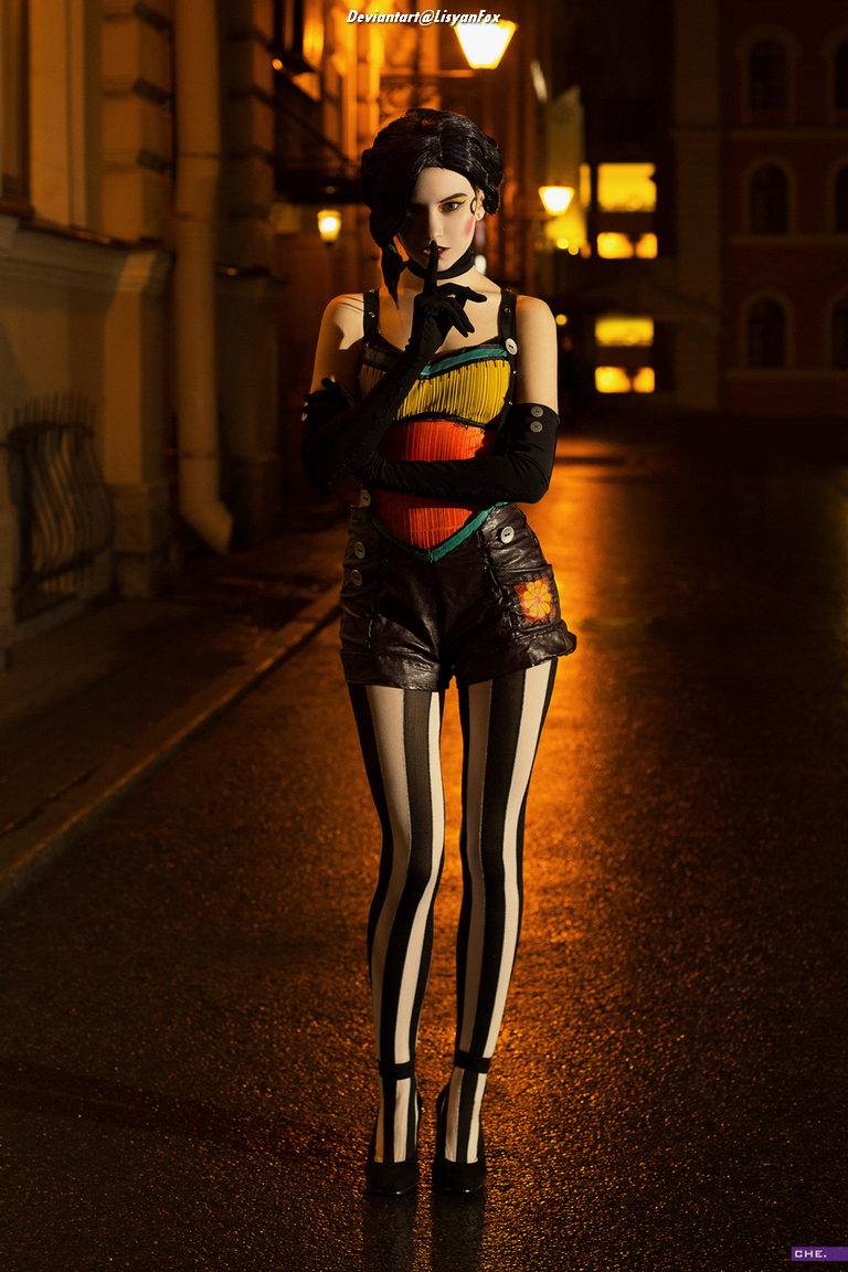A person standing on a wet street at night, wearing a colorful top, shorts, and striped tights, with a finger raised to their lips as a gesture of silence.