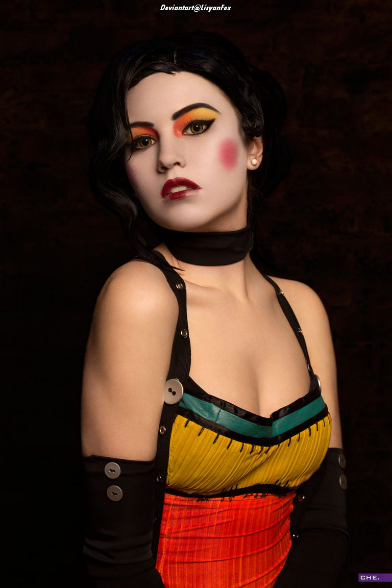 A woman with bold makeup, including colorful eyeshadow and red lips, poses in a vibrant dress with a yellow, green, and orange pattern, against a dark background.