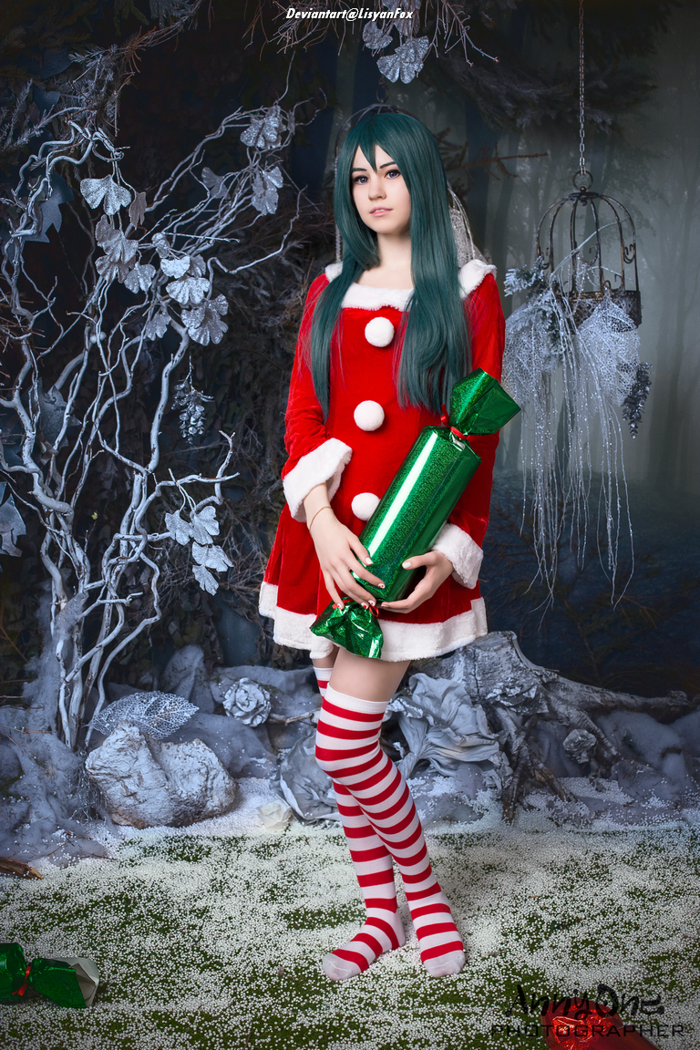 A person dressed as a holiday character in a red and white outfit, holding a green festive cracker, standing in a winter-themed setting with snow and decorative elements.
