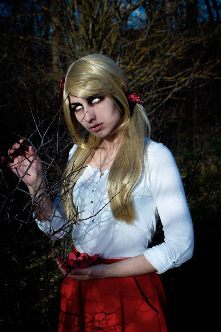 A young woman with long blonde hair and dark eyes stands in a forest, wearing a white top and a red skirt, holding a flower and reaching for branches.