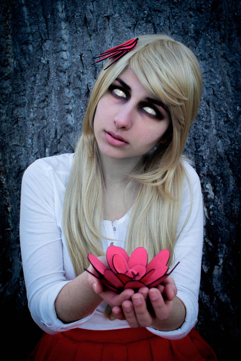 A young woman with long blonde hair and a white shirt holds a pink paper flower, looking upwards with pale eyes and a neutral expression, set against a textured tree bark background.