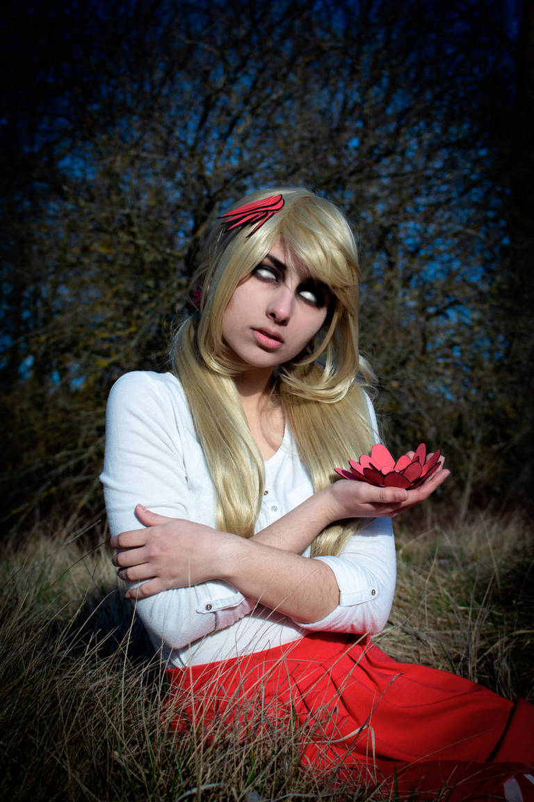 A person with long blonde hair, wearing a white sweater and a red skirt, sits in a grassy area holding a red paper flower, with a serious expression and unique eye makeup.