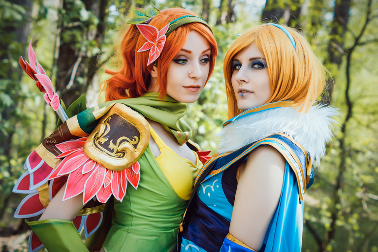 Two women in vibrant fantasy costumes stand close together in a forest setting, one dressed in green with floral elements and the other in blue and white with fur accents.