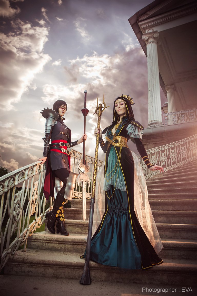 Two individuals in elaborate costumes standing on a staircase. One is dressed in armor with a dark color scheme, holding a staff, while the other wears a regal outfit with a long train and a crown, with a staff adorned with symbols. The background features dramatic clouds and architectural details.