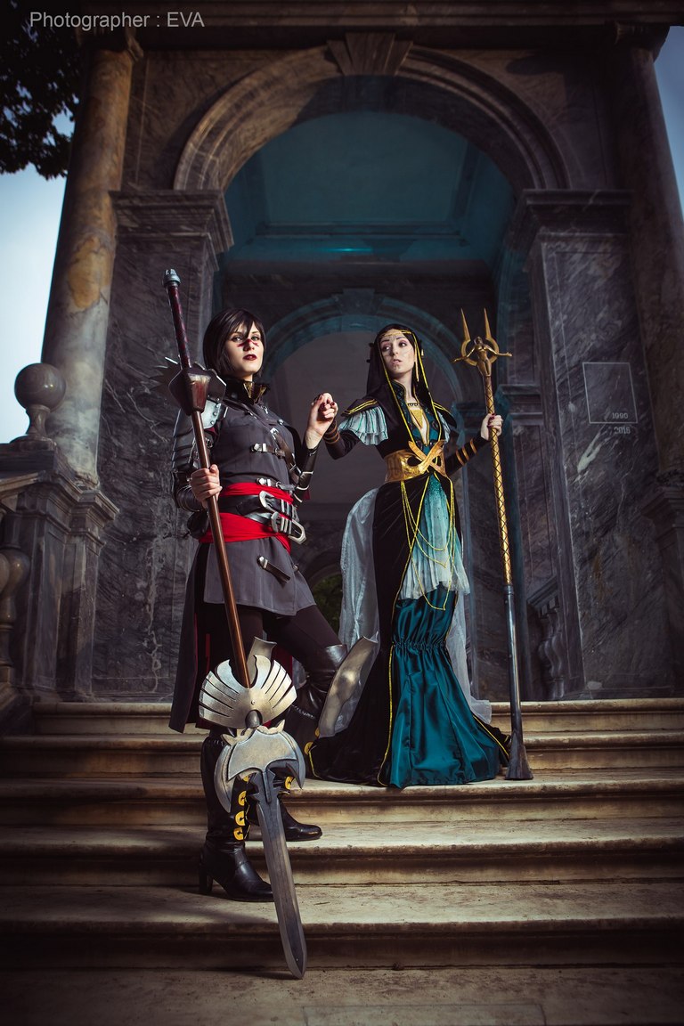 A scene featuring two individuals in elaborate fantasy cosplay outfits. One character holds a sword and staff, while the other adorns a flowing gown with a large staff, set in a grand staircase environment.