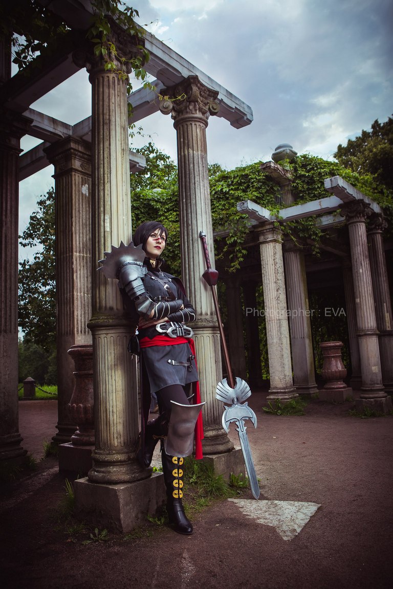 A person in a dark fantasy costume leaning against a pillar in a classical outdoor setting, with cloudy skies in the background.