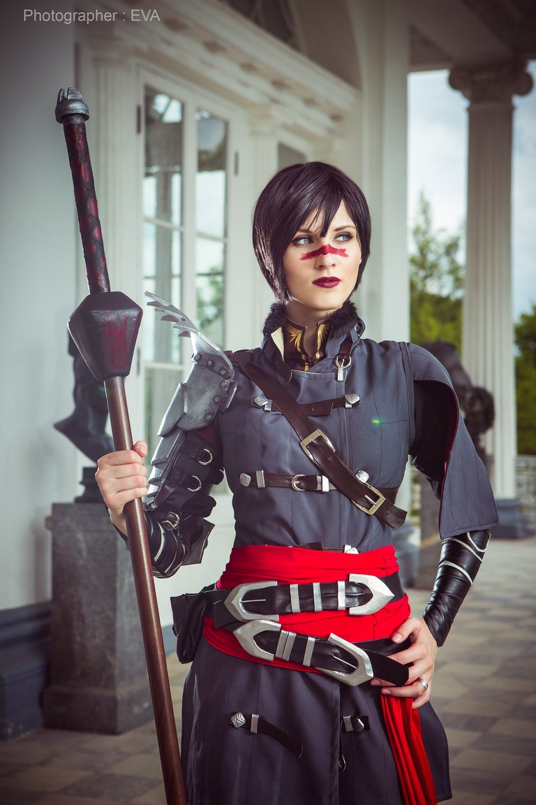 A person in a dark costume with silver armor details and a red sash, holding a staff, posed in an outdoor setting with architectural elements.