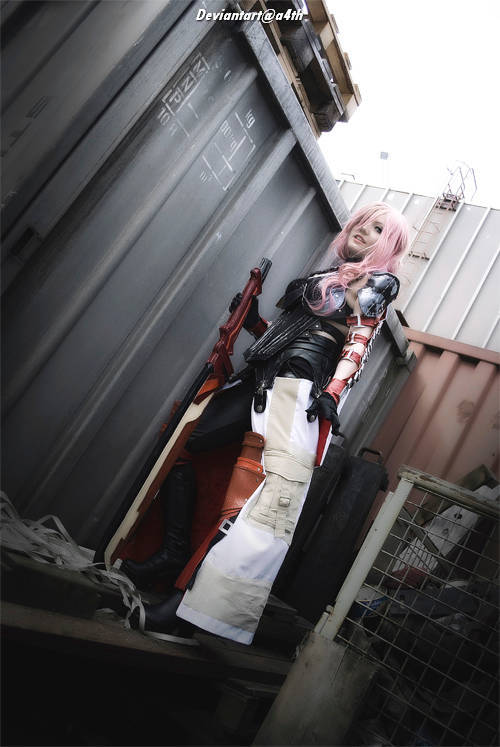 A person in a striking costume with pink hair stands next to a shipping container, holding a weapon.