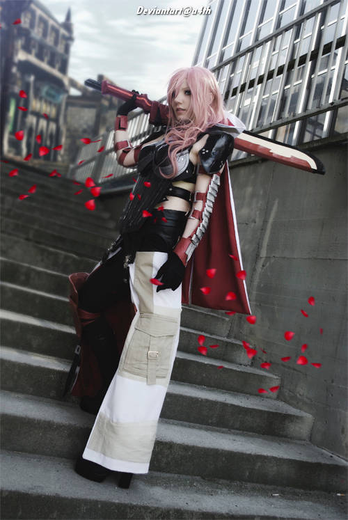 A person in a striking cosplay outfit, featuring pink hair and a dramatic red cloak, poses on a set of stairs surrounded by scattered rose petals.