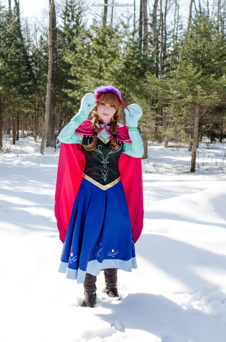 A person dressed in a colorful winter costume inspired by a princess character, standing in a snowy forest. The outfit features a pink cape, blue skirt, and intricate designs, with trees in the background.