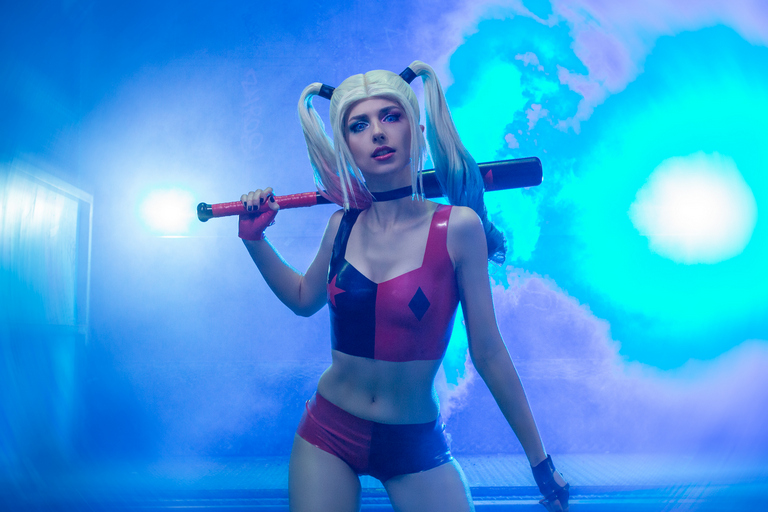 A person dressed as a character with blonde pigtails, wearing a red and black outfit, holding a baseball bat, posed against a blue smoky background.