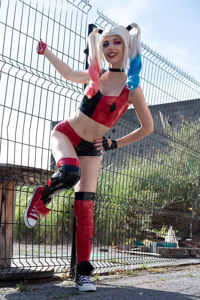 A person posing in a colorful costume with red and black elements, standing by a fence with a playful expression.