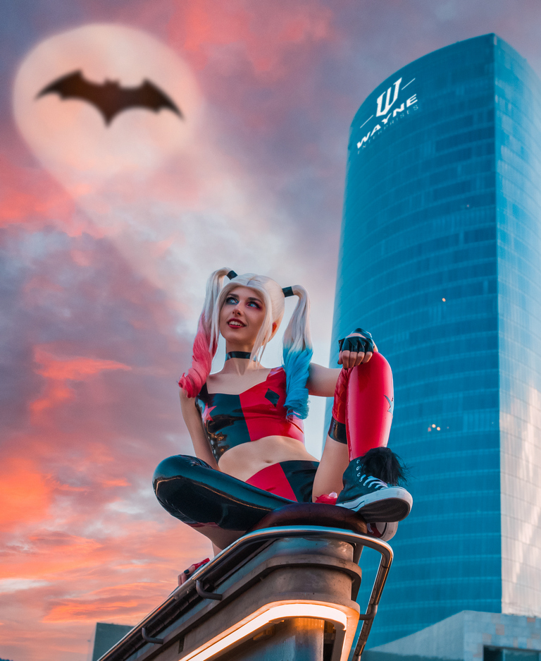 A person dressed as Harley Quinn sits on a railing with a skyscraper in the background, featuring a Batman symbol in the sky and a colorful sunset.