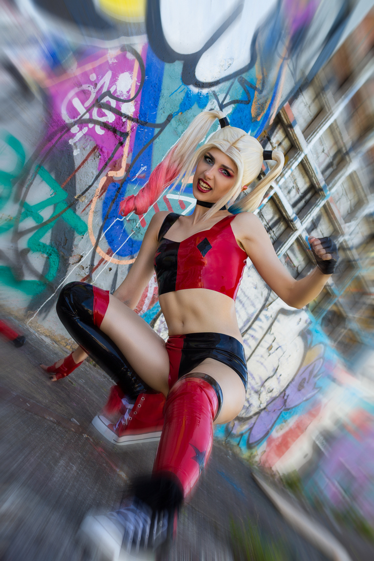 A person in a playful and dynamic pose, dressed in a red and black superhero costume with a cropped top and thigh-high boots, against a colorful graffiti background.