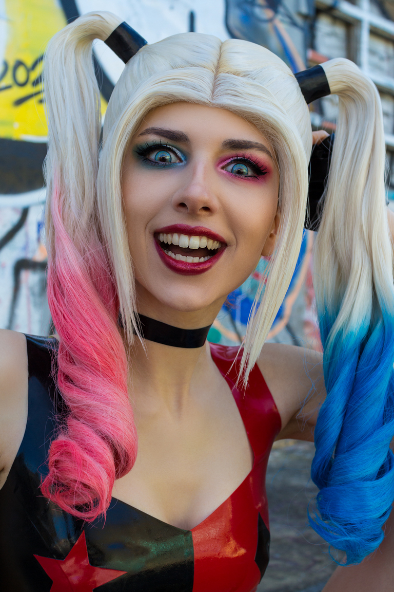 A person with multicolored hair styled in pigtails, wearing a colorful outfit, and posing enthusiastically in front of a graffiti-covered wall.