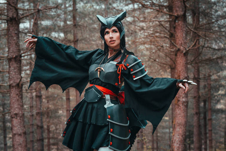 A person dressed in a bat-themed costume with large sleeves, standing in a forest setting with trees in the background.