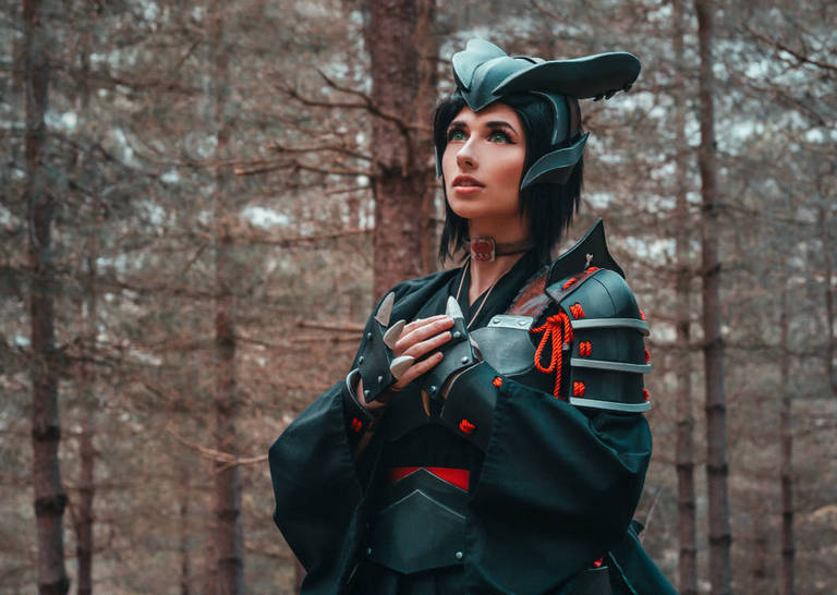 A person in a detailed black cosplay outfit with armor elements, standing in a forest setting, looking up thoughtfully.