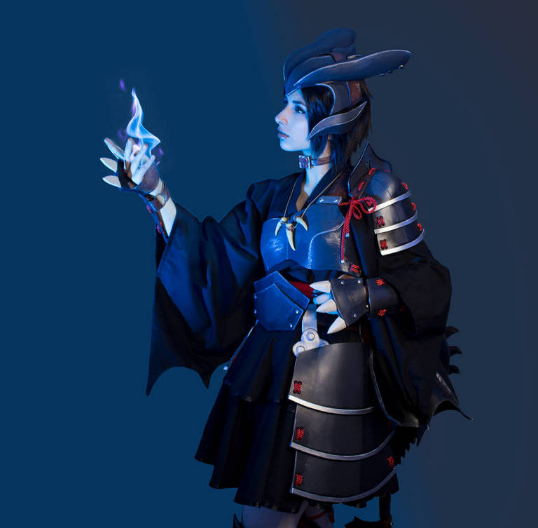 A person in a dark outfit holds a glowing blue flame in their hand, showcasing a detailed costume with armor and a flowing cape.