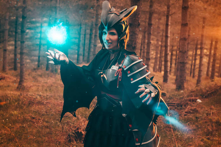 A person in a dark, ornate costume with a bat-like design, wielding a glowing blue orb in a forest setting.