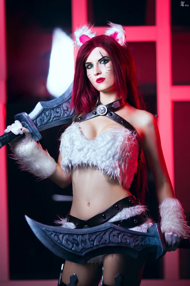 A cosplayer dressed as a character with cat-like features, wearing a fluffy white outfit and holding twin swords. The background is illuminated with red lighting.