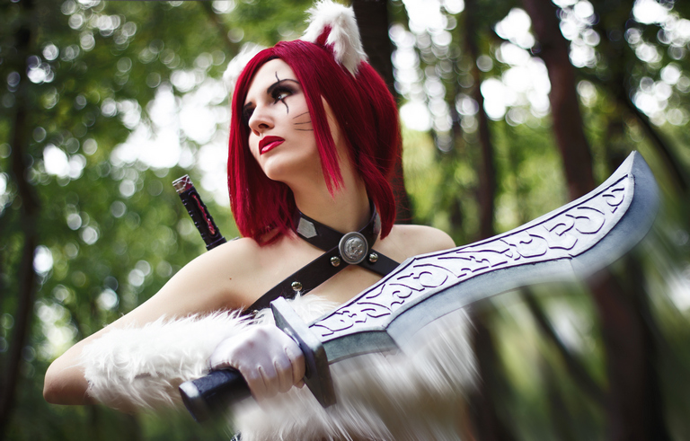 A woman in a forest setting, wearing a cosplay outfit with red hair, cat-like ears, and holding a large sword.
