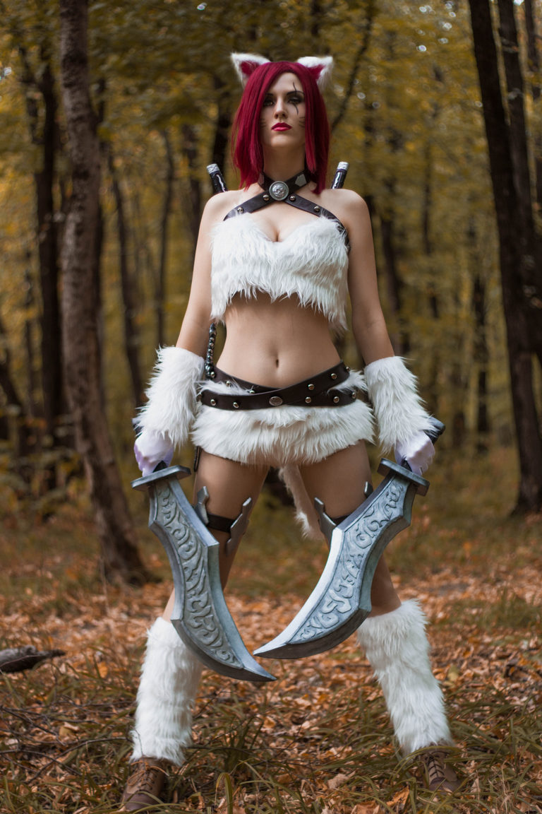A person dressed in a fantasy warrior costume with cat-like ears, holding dual curved swords in a forest setting during autumn.