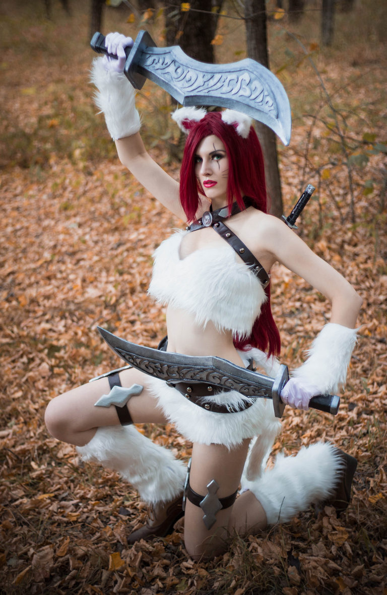 A woman in a fantasy costume with red hair and animal ears, holding two swords, kneeling in a forest with autumn leaves.