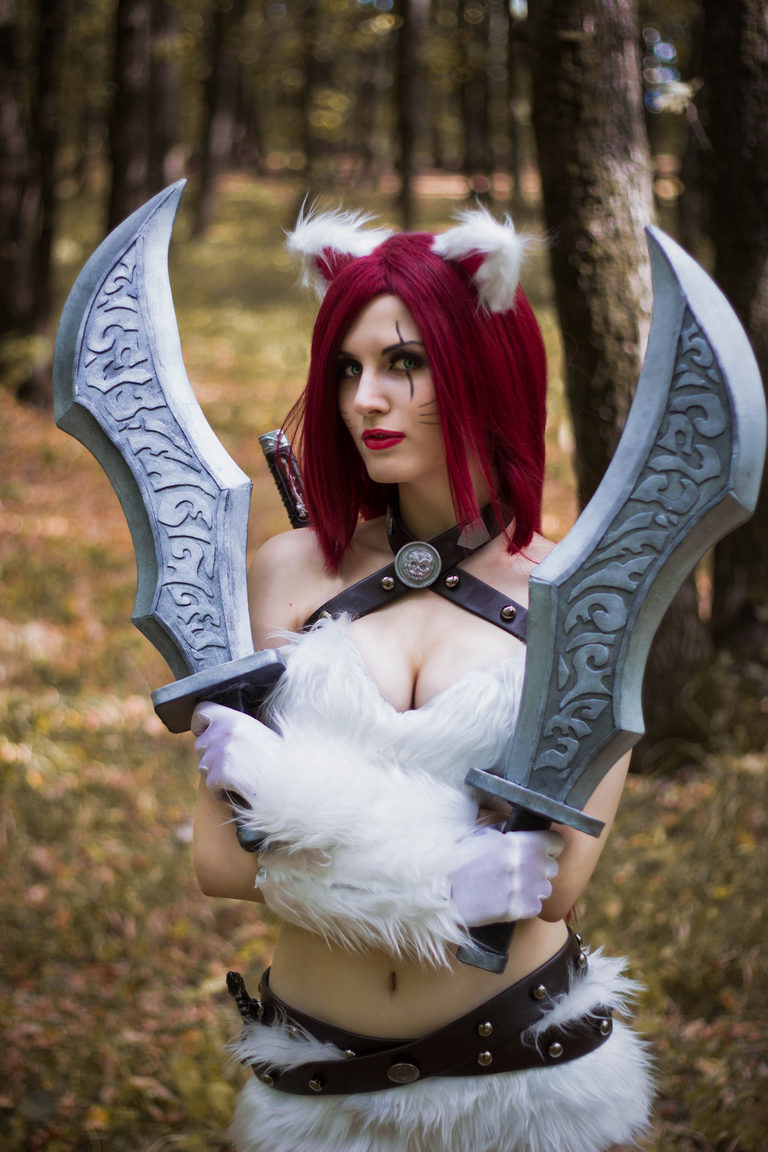 A person wearing a furry white outfit with cat ears and holding two large decorated swords, posing in a forest setting.
