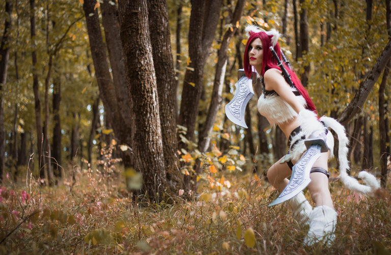 A person in a fantasy-themed costume with red hair poses in a forest, surrounded by autumn foliage.
