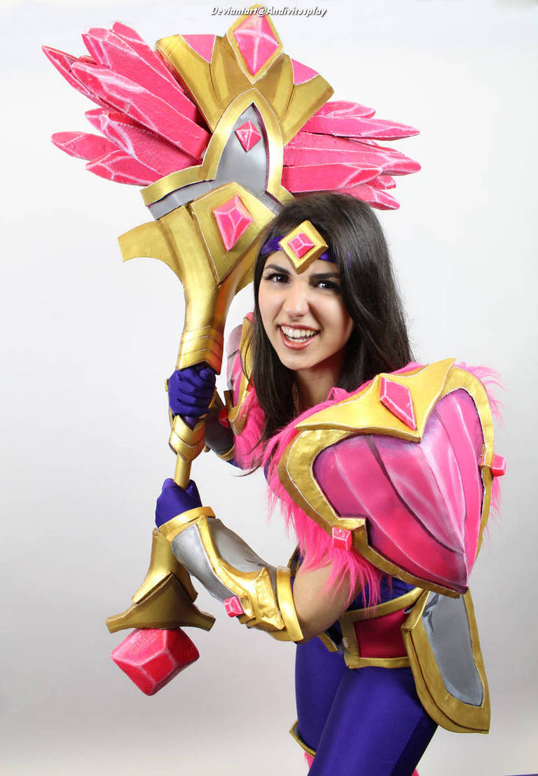 A person in a vibrant cosplay costume featuring pink and gold armor, holding a large, decorative staff with bright crystals, and smiling at the camera.