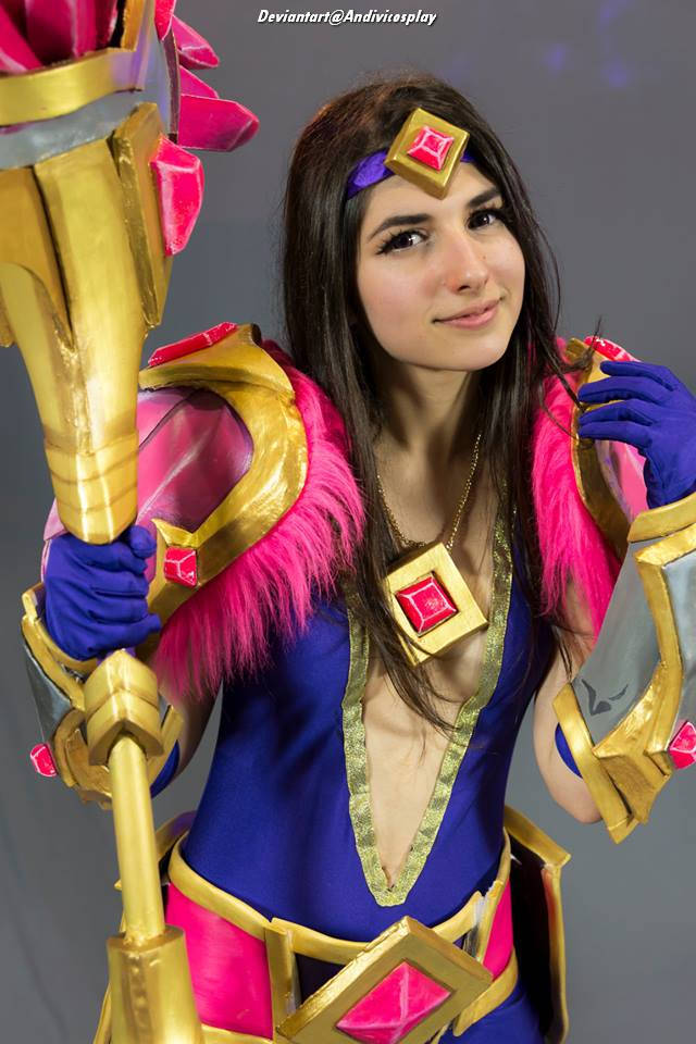 A person wearing a vibrant costume with pink and gold accents, posing with a large staff adorned with pink gems.