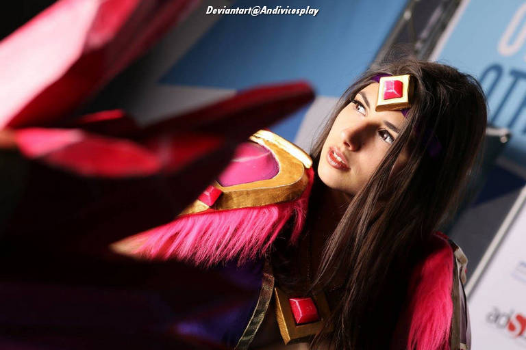 A close-up photo of a woman in a vibrant costume with pink feathers and gold accents, posing thoughtfully. The background features blurred colors, suggesting a convention or staged setting.