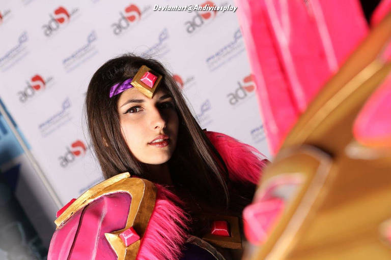 A person dressed in a colorful cosplay costume with pink and gold accents, showcasing long dark hair and a headpiece with a gem, posing against a branded backdrop.