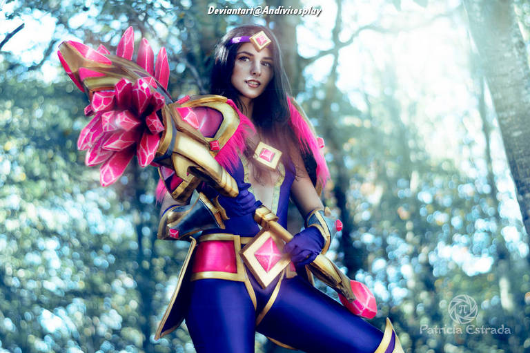Woman cosplaying as a character with a large, ornate staff adorned with pink and gold accents, set in a forest background with soft, natural lighting.