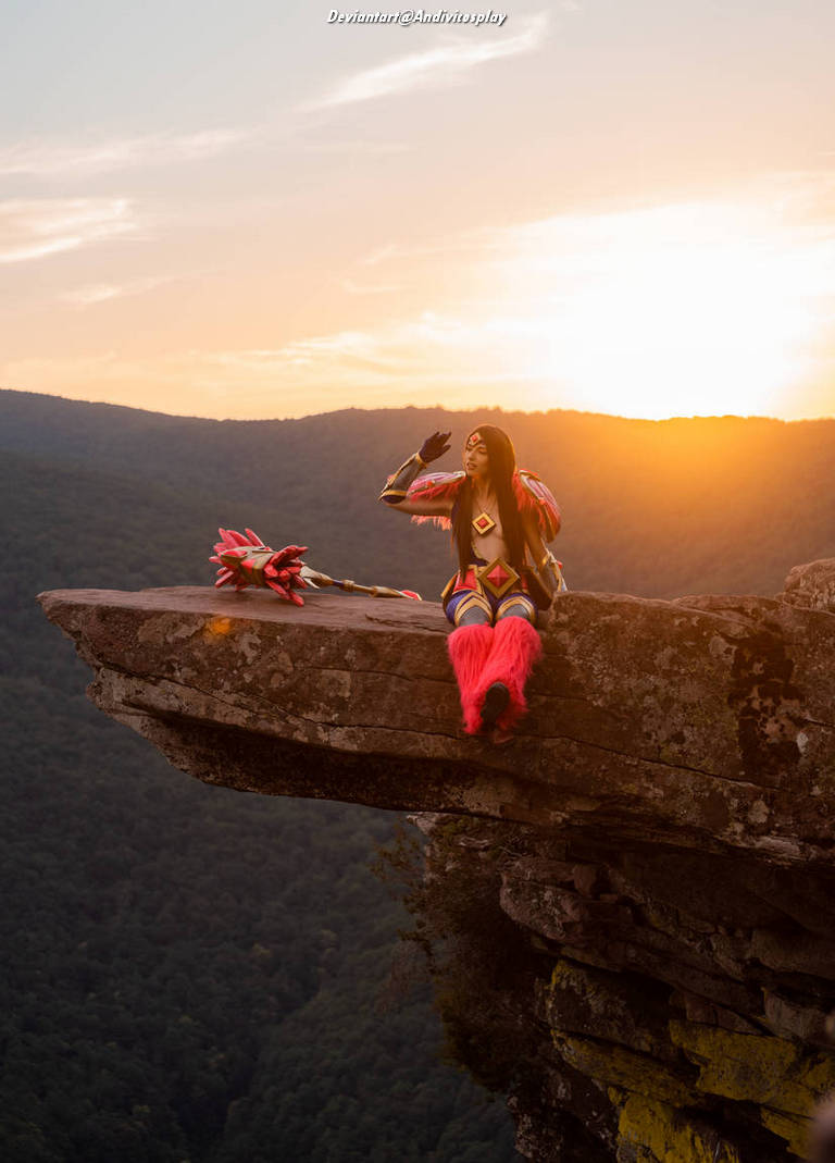 A person dressed in colorful cosplay attire, featuring bright fur and geometric patterns, sitting on a rock ledge with a scenic mountainous background during sunset.