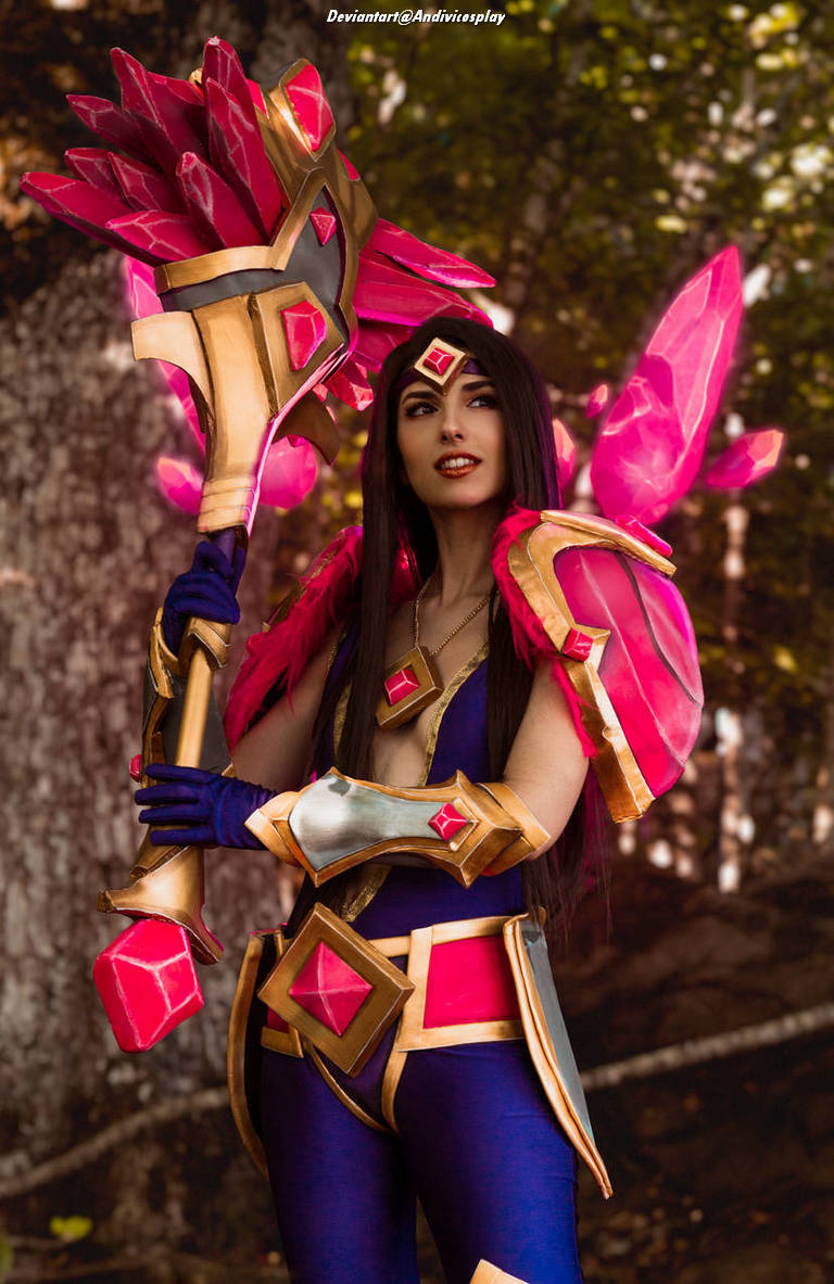 A person in a colorful fantasy costume featuring pink and gold elements, holding a large staff adorned with crystals. The background is a blurred natural setting.