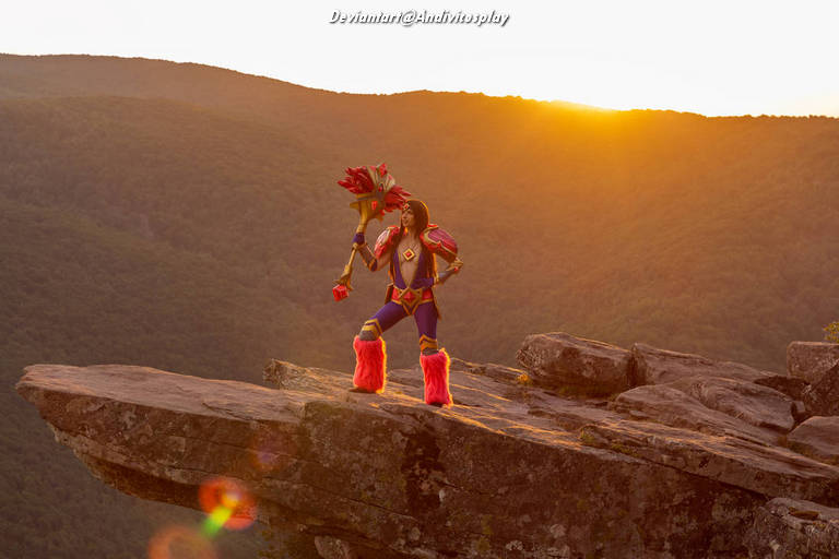 Person in a colorful superhero costume stands on a rocky ledge, holding a staff with a red flower, against a sunset backdrop over a mountainous landscape.