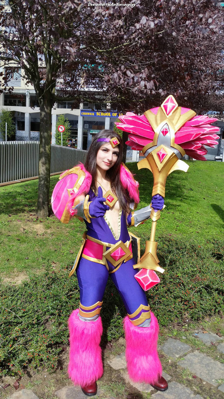 A cosplayer dressed as a character with vibrant pink and purple attire, featuring fluffy pink leg warmers and holding a decorative staff in a park setting.