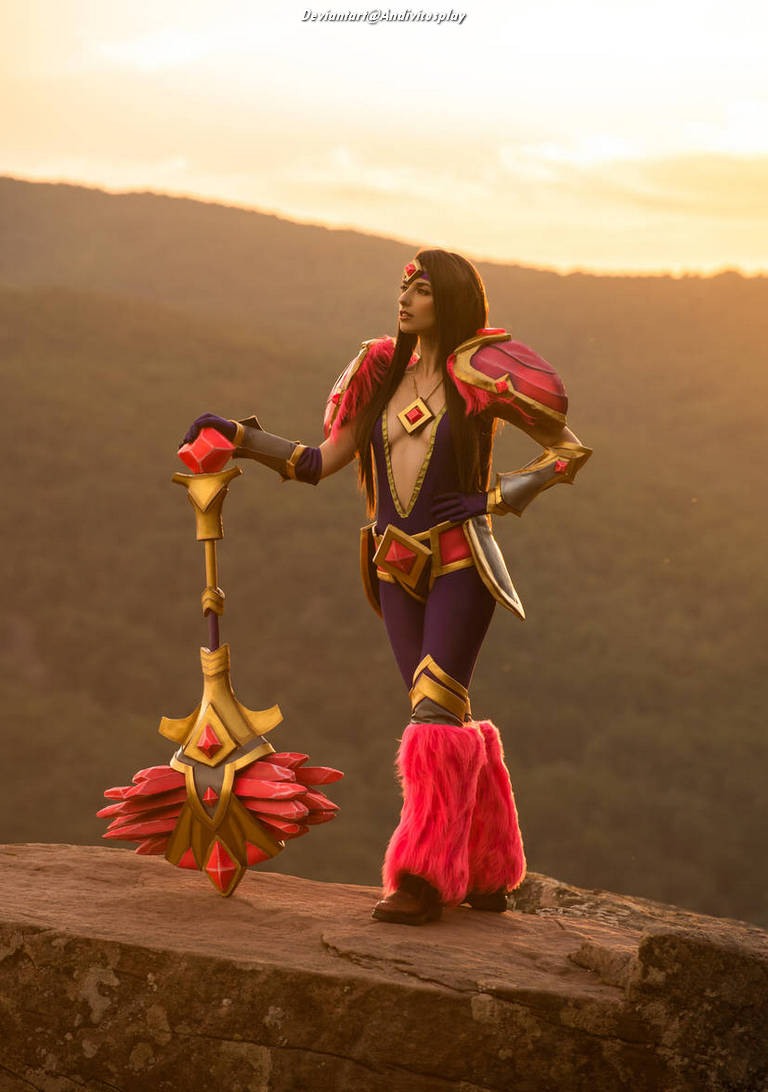 A cosplayer dressed as a fantasy character stands confidently on a rocky ledge at sunset, wearing vibrant purple attire with pink fur accents and holding a large ornate staff.