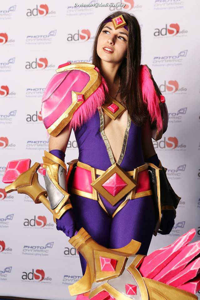 A young woman in a vibrant costume inspired by a fantasy character, featuring purple and pink armor with decorative elements, posing confidently in front of a backdrop with photography studio logos.