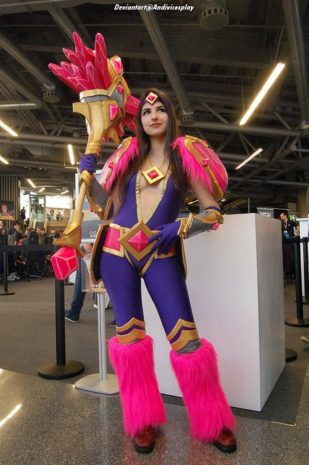 A person in a vibrant purple and pink costume with ornate armor and accessories, posing confidently at a convention. They are holding a staff adorned with jewels.
