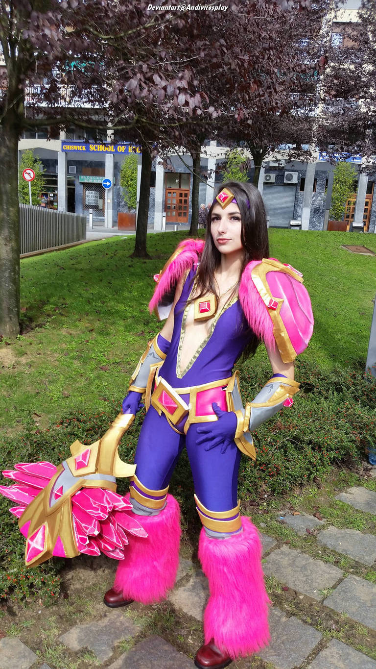 A cosplayer dressed as a fantasy character, wearing a purple and pink costume with extravagant armor and a large weapon, standing in a park with trees and a building in the background.