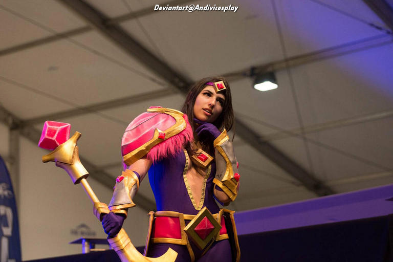 A cosplayer dressed as a character from a fantasy series, featuring a colorful costume with pink and gold accents, posing thoughtfully on stage.