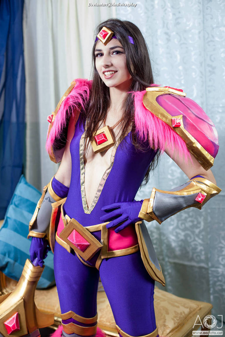A woman in a colorful fantasy costume with pink and purple accents poses confidently with one hand on her hip. She has long dark hair and is smiling, set against a backdrop of soft drapes and cushions.