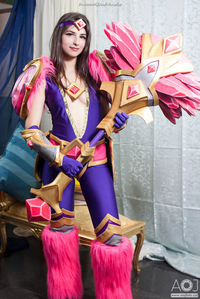A cosplayer dressed in elaborate fantasy armor featuring purple, pink, and gold colors, holding a large decorative weapon, with fluffy pink accents on her outfit and boots.
