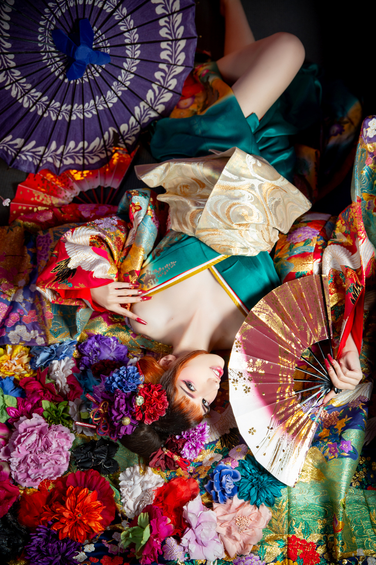 A woman dressed in a colorful kimono poses amid vibrant flowers and traditional decor, holding a fan and surrounded by parasols.